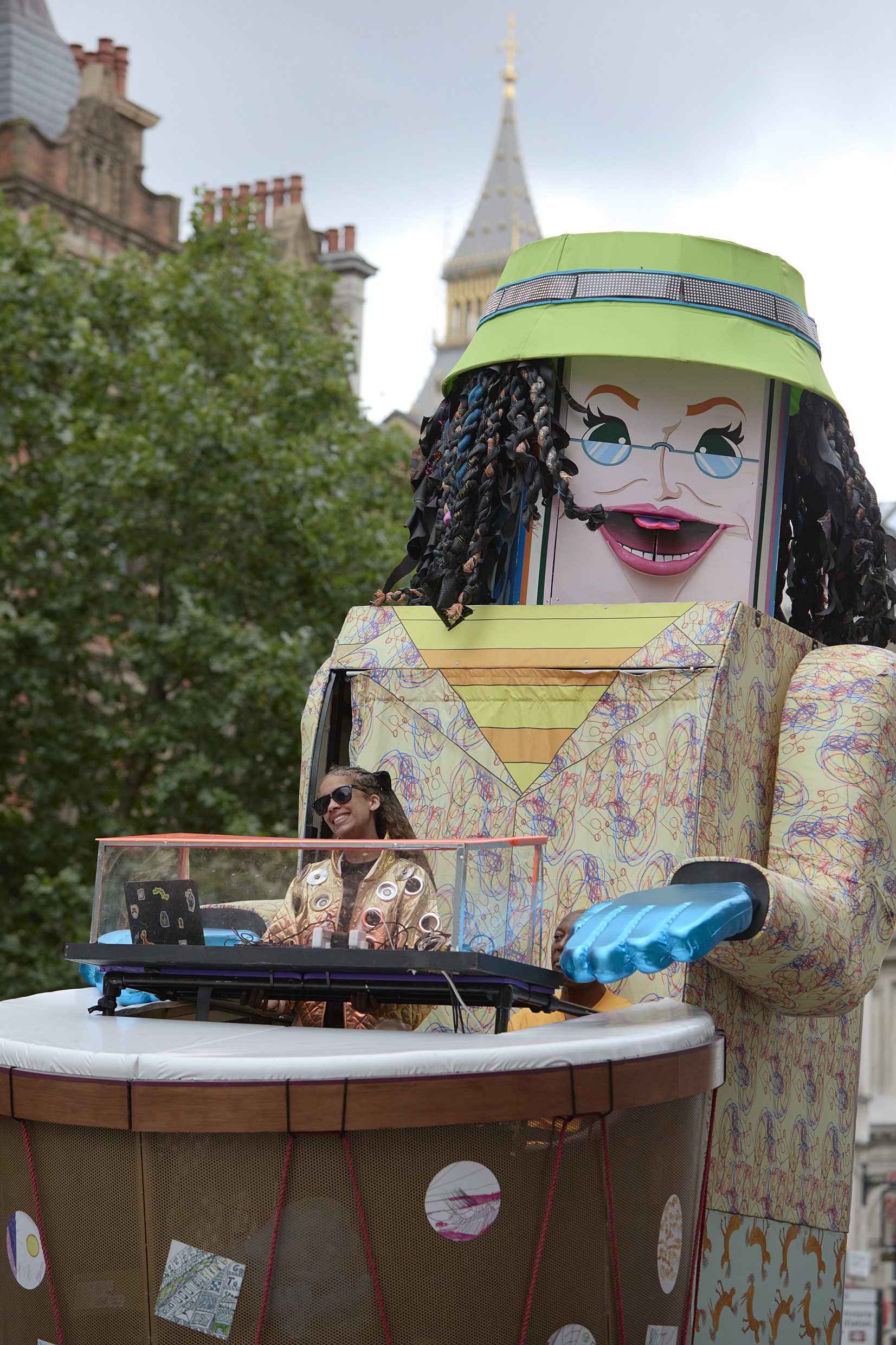 A giant puppet with a green hat and black hair, with a DJ set in the foreground.