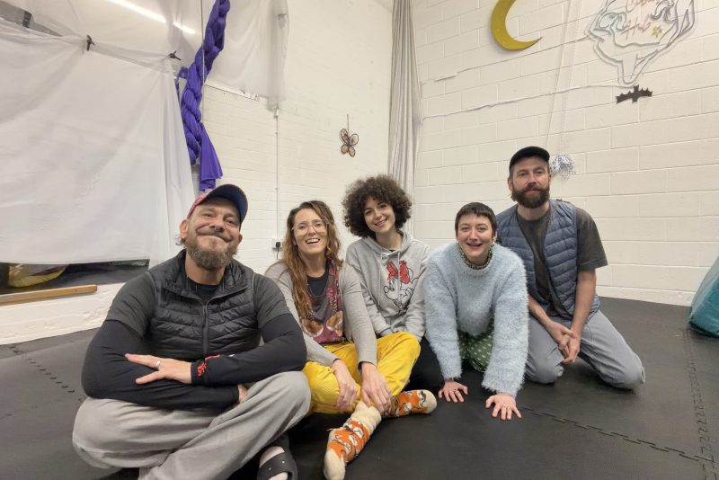 An image of 3 women and 2 men sat on the floor in an art studio facing the camera.