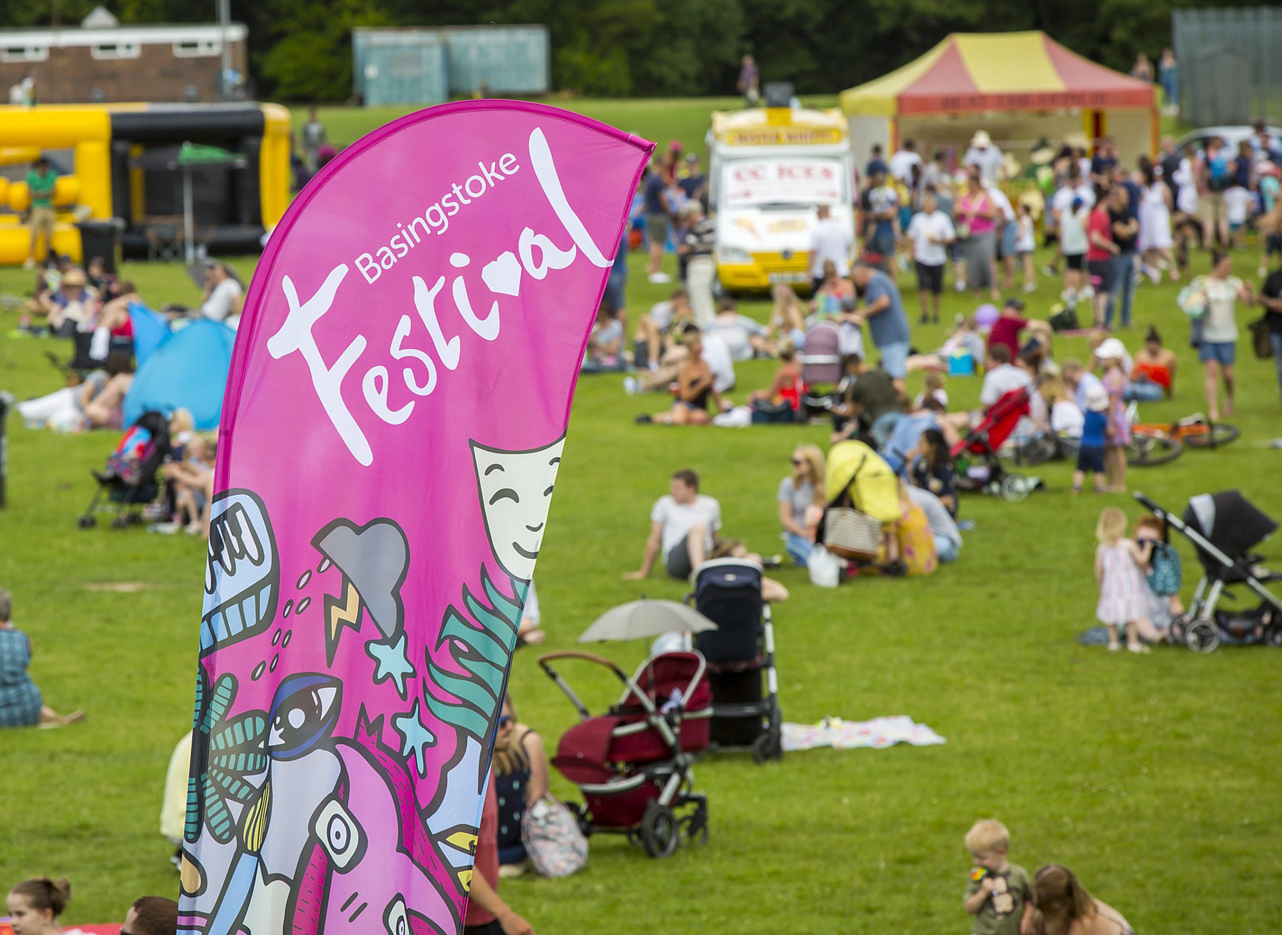 An image of a Basingstoke Festival banner in the foreground with a crowd sat on a large patch of grass in the background.
