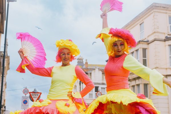 2026 Touring Network Partners. An image of two female performers wearing bright and colourful orange and yellow dresses, wigs and are holding pink and white fans.