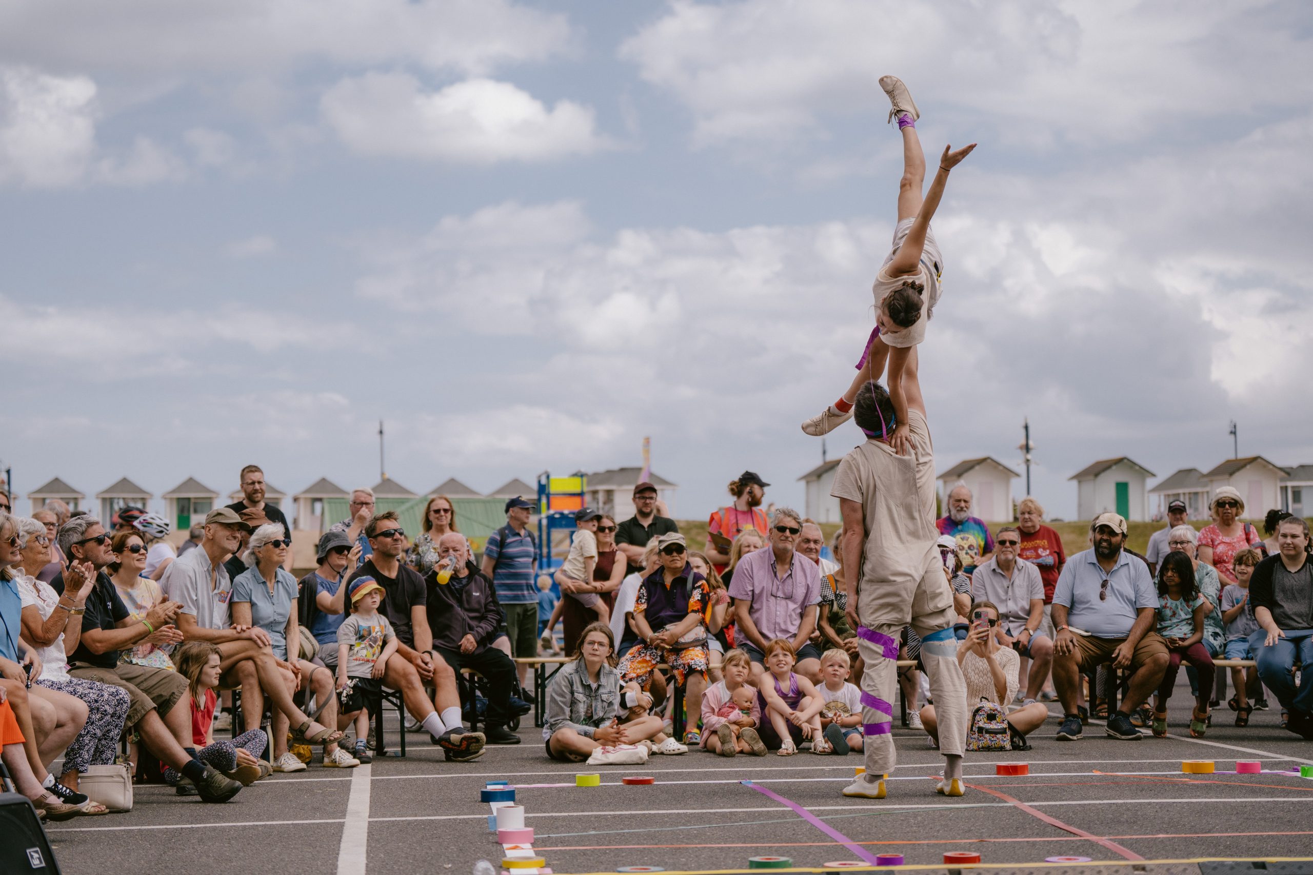 An image of two performers. The male performer is lifting up a female performer into the air, and there is a crowd stood and sat down behind them watching.