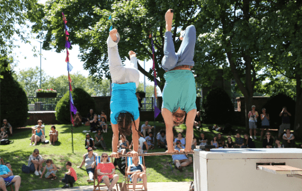An image of two people doing a hand stand on top of a horizontal suspended pole with trees in the background.