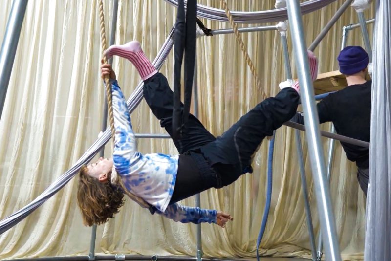 An image of a female performer hanging from a rope with a cream curtain in the background.