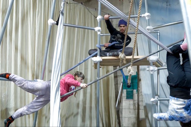 An image of three performers balancing on and hanging from a steel structure with a cream curtain in the background.